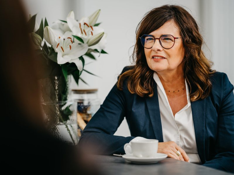 Beraterin im Business-Outfit im Gespräch, mit Kaffeetasse am Tisch.